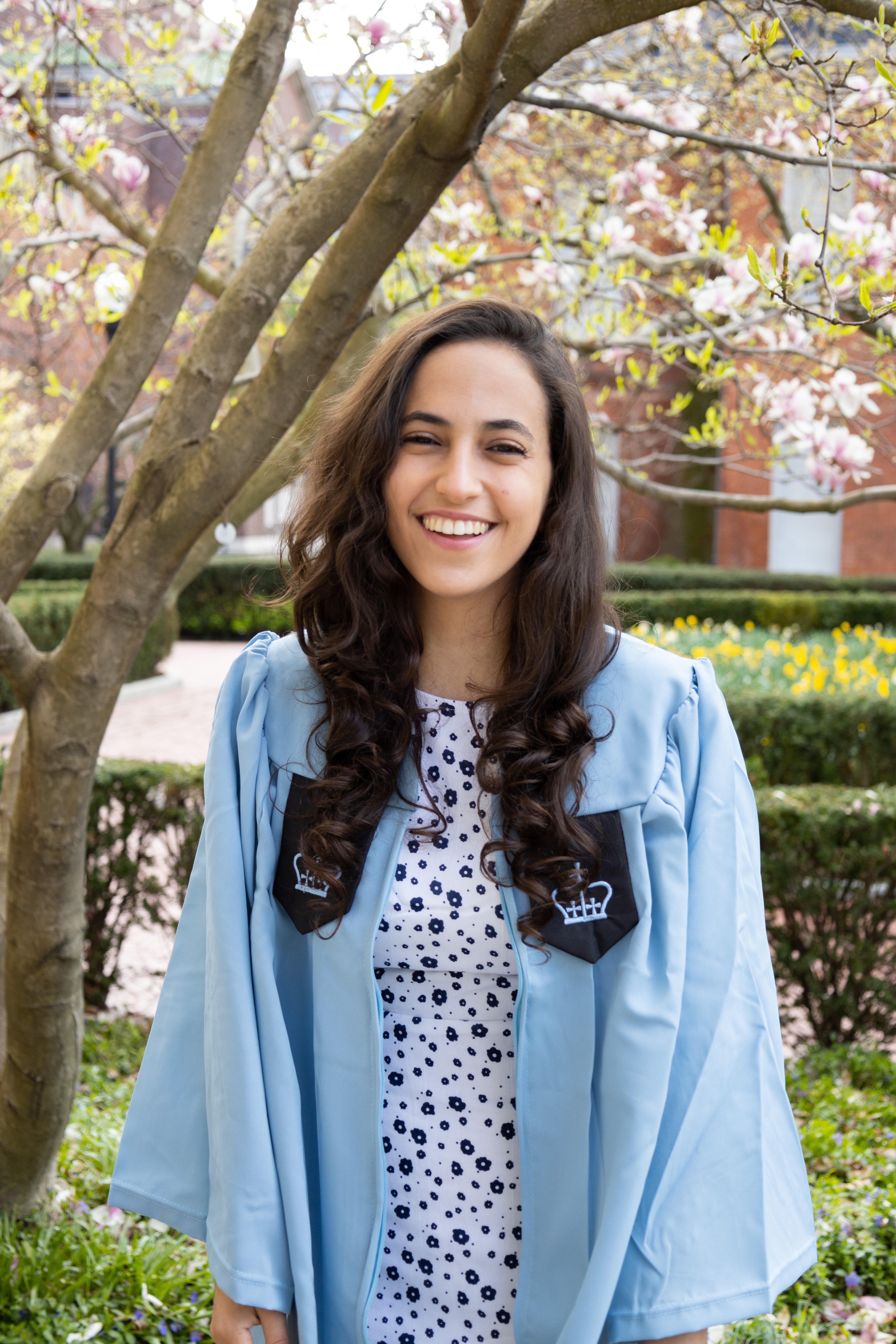 Sophia Houdaigui in her light blue graduation gown standing outside in front of a tree
